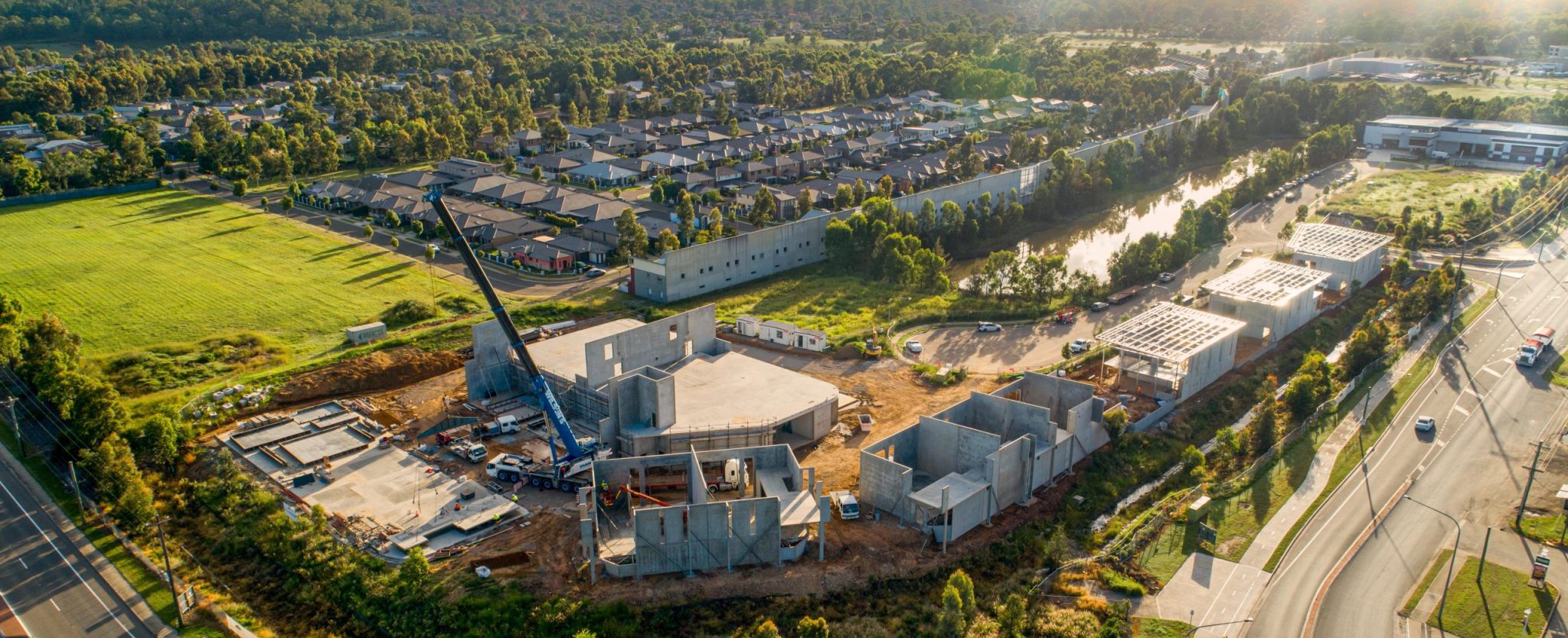 Aerial view of a construction site with several partially built concrete structures and a blue crane lifting materials, surrounded by roads, green fields, residential houses, and trees in the background.