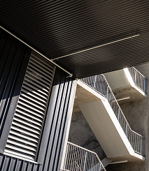 Architectural view of a modern building corner featuring vertical metal siding and an outdoor concrete staircase with metal railings, highlighted by natural light and shadows.