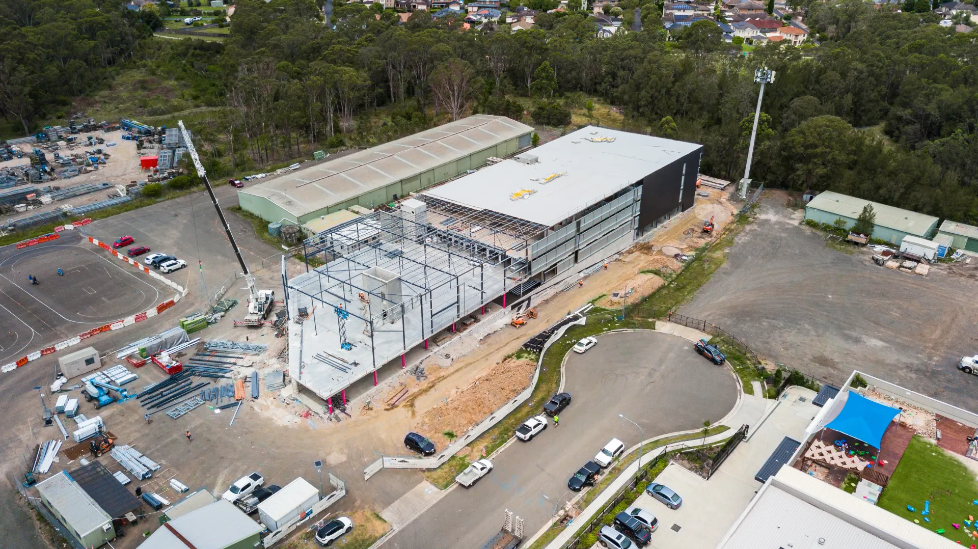Aerial view of a construction site with a large building partially completed, surrounded by vehicles, construction materials, and equipment, with a wooded area and residential houses in the background.