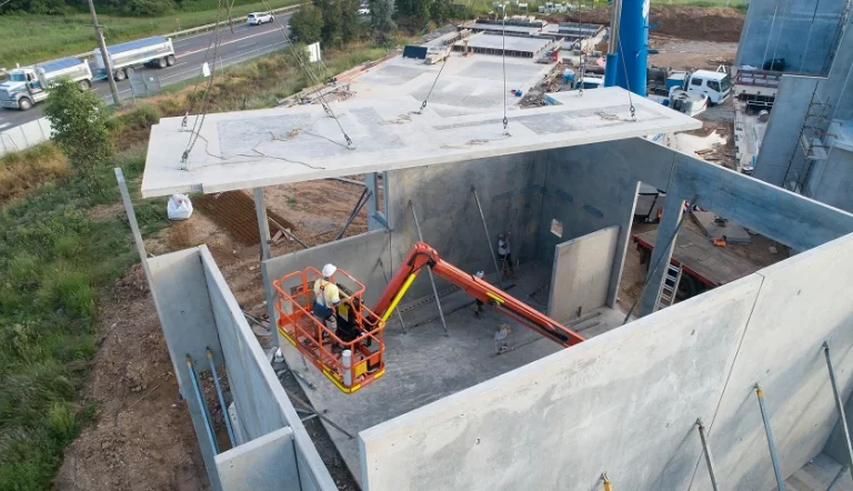 Construction site with two workers in an orange boom lift installing a large concrete panel above a partially built concrete structure next to a road with passing vehicles.
