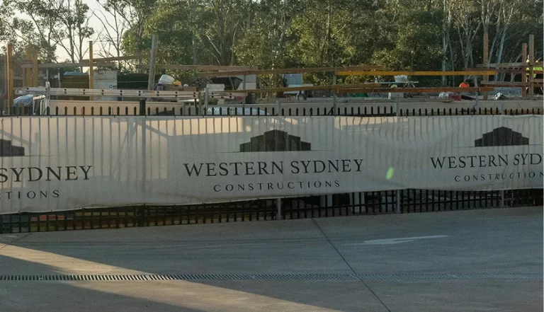Construction site with a black metal fence and white banners displaying the logo and text 'Western Sydney Constructions' in front of a partially built structure surrounded by trees.
