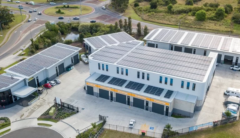 Aerial view of a commercial complex with multiple buildings featuring solar panels on their roofs, surrounded by roads, parking areas with cars, and grassy fields.