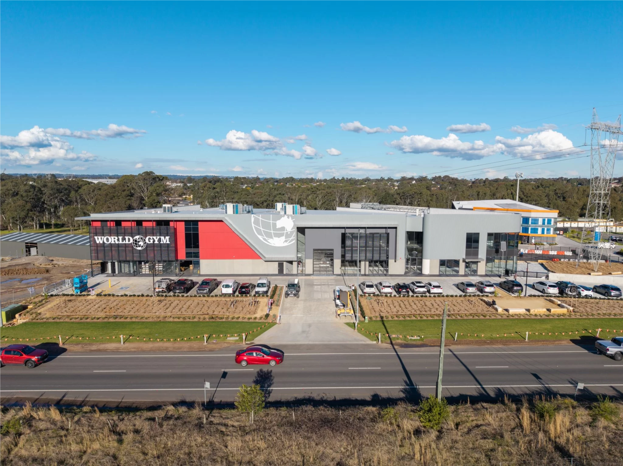 Large modern building with WORLD GYM signage on the left side, featuring red, black, and gray exterior walls, parking lots with cars on both sides, and a road with moving vehicles in front under a clear blue sky.