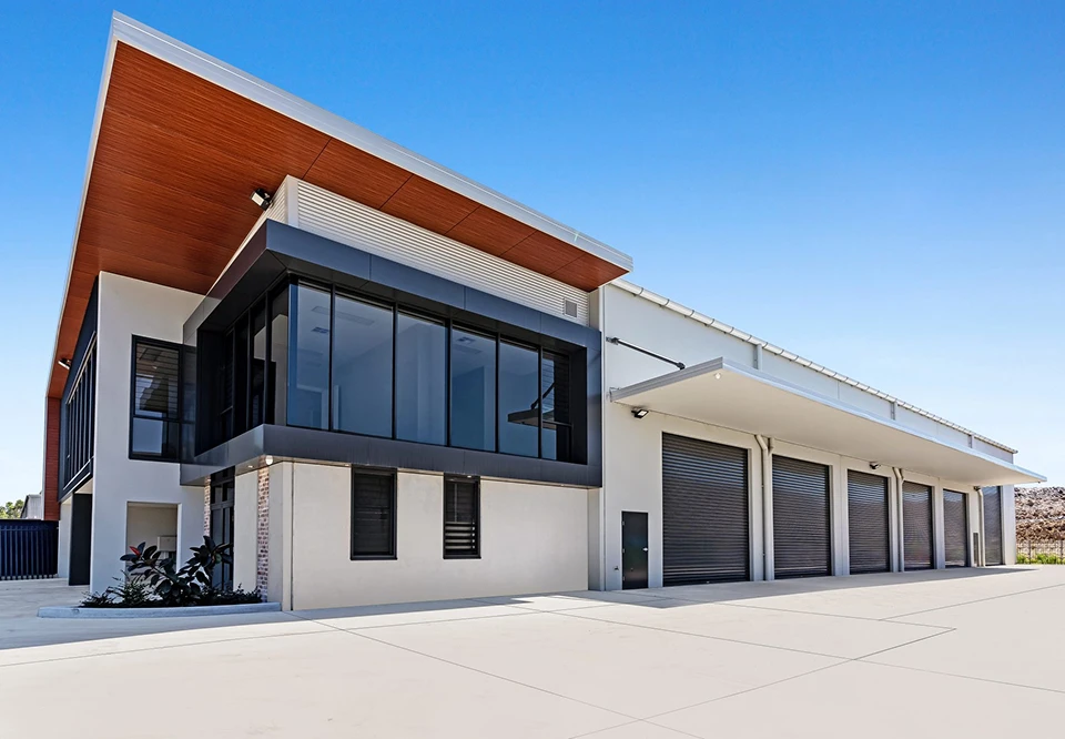Modern industrial building with large black-framed glass windows, white walls, a slanted wooden ceiling overhang, and multiple closed black garage doors along a concrete driveway under a clear blue sky.