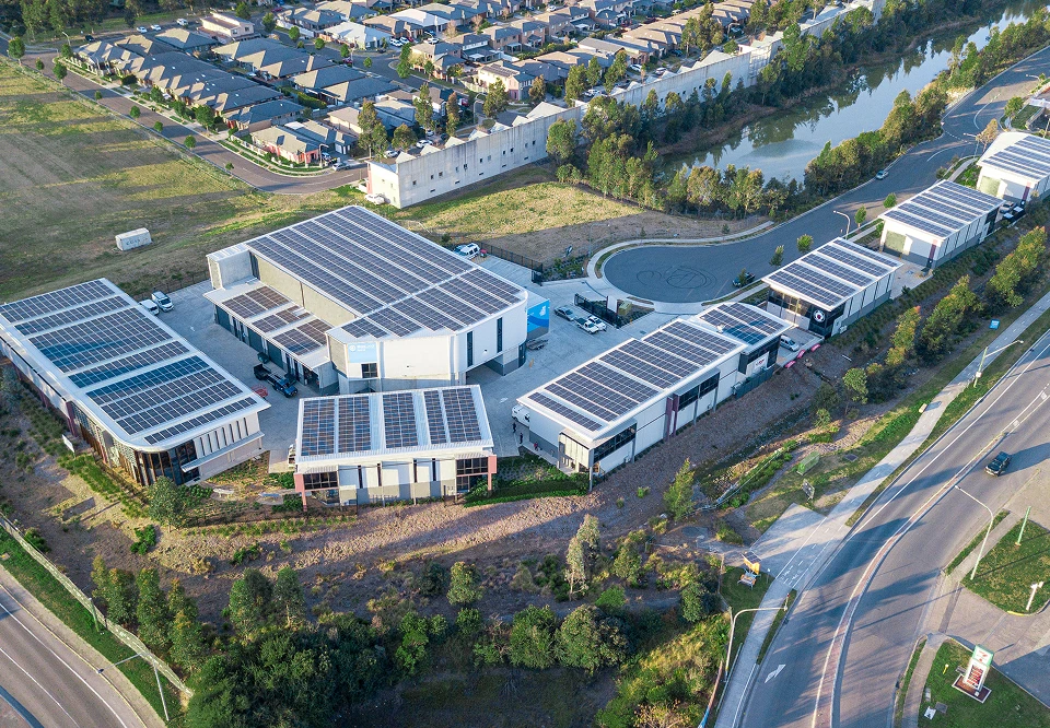 Aerial view of a modern business park with multiple buildings featuring solar panels on their roofs, surrounded by roads, trees, a small body of water, and residential houses in the background.