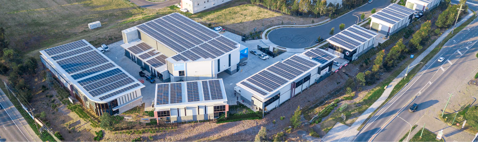 Aerial view of a business complex with multiple buildings equipped with solar panels on their roofs, surrounded by roads and greenery.