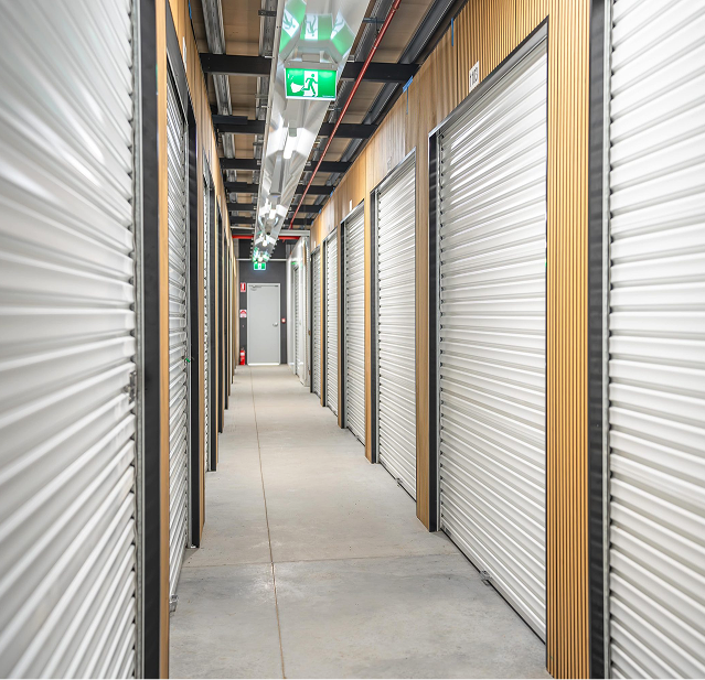Indoor corridor with closed silver roll-up storage unit doors on both sides and a gray exit door at the end, illuminated by overhead lights and green exit signs.