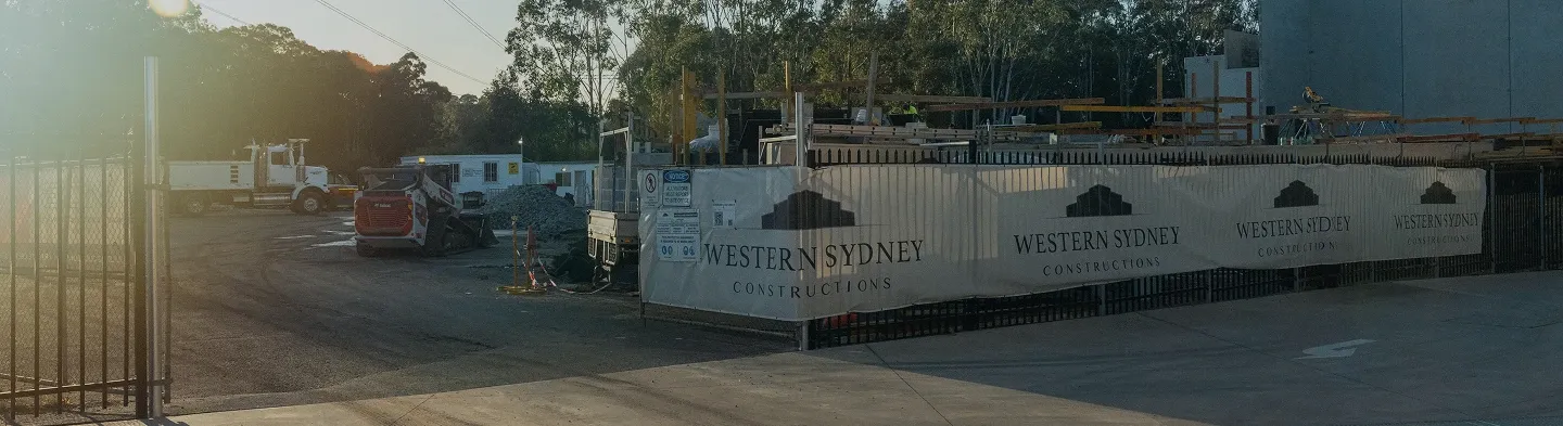 Construction site fenced with banners reading 'Western Sydney Constructions,' featuring construction materials, a white truck, and a mini excavator under late afternoon light.