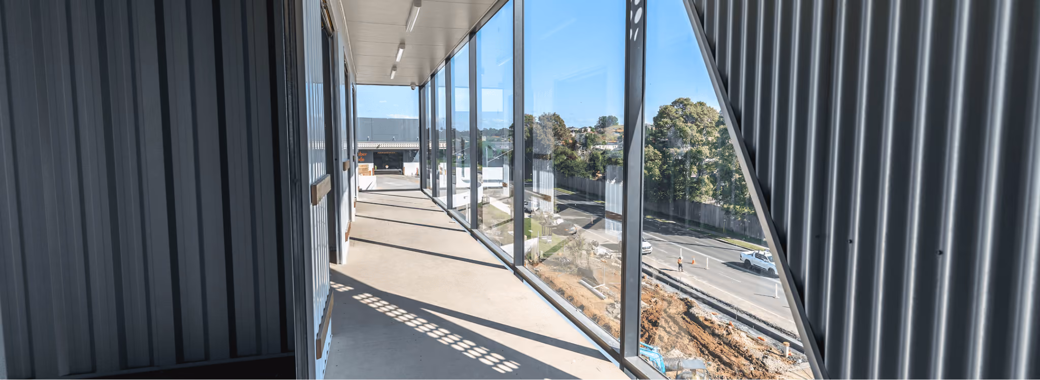 Sunlit modern balcony with large glass windows on the right and dark corrugated metal walls on the left, overlooking a street and some trees.