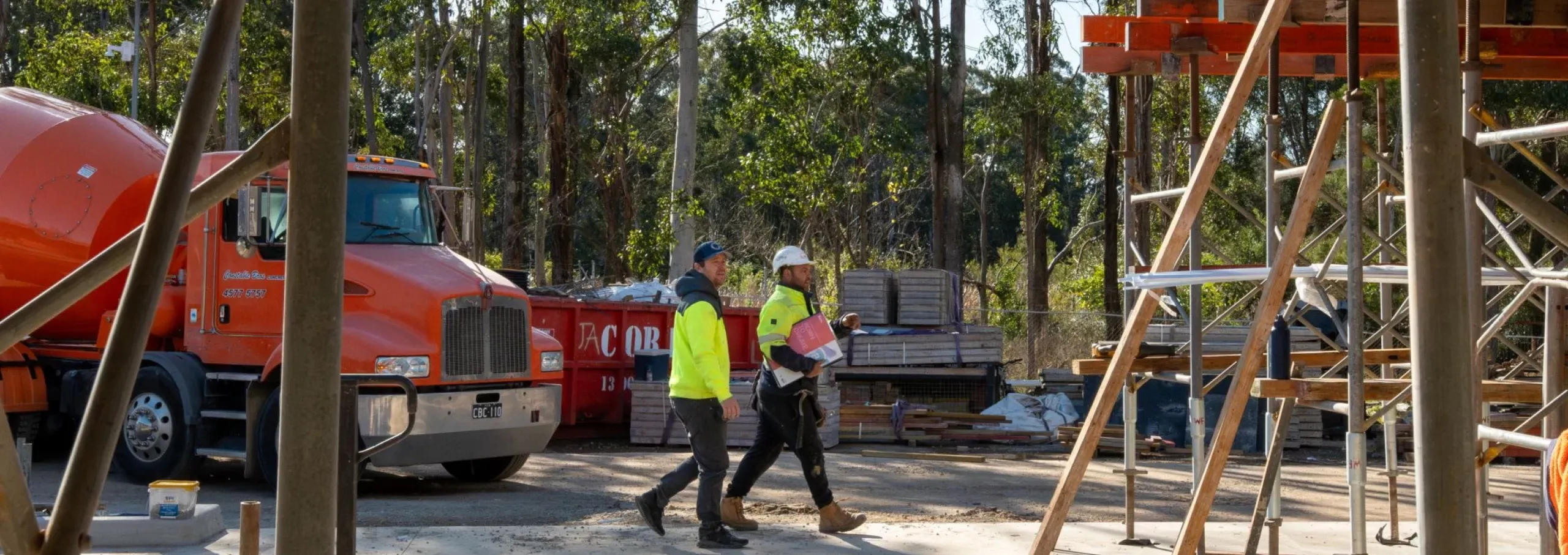 Two construction workers in high-visibility jackets and helmets walking on a construction site near scaffolding and an orange cement mixer truck with trees in the background.