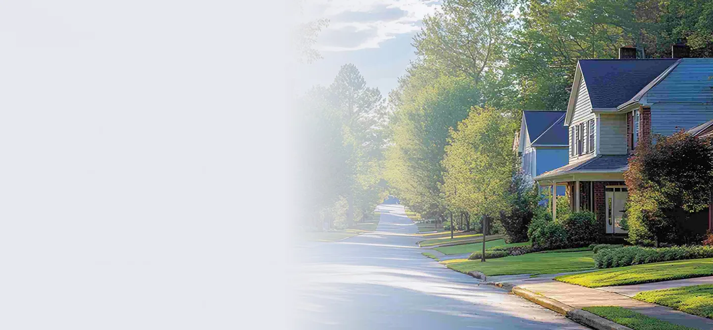 Quiet suburban street lined with green trees and houses under a partly cloudy sky.