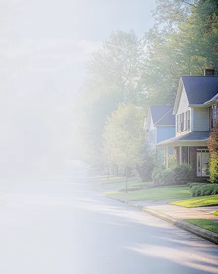 Quiet suburban street with houses, green lawns, trees, and soft sunlight.