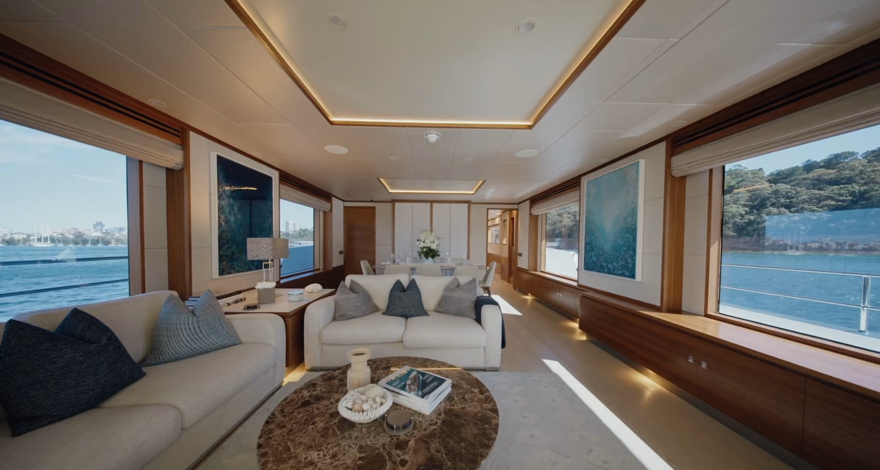 Luxurious yacht interior featuring spacious lounge area with two beige sofas, round marble coffee table with decorative items, large windows showing water and greenery outside, and modern ceiling lighting.