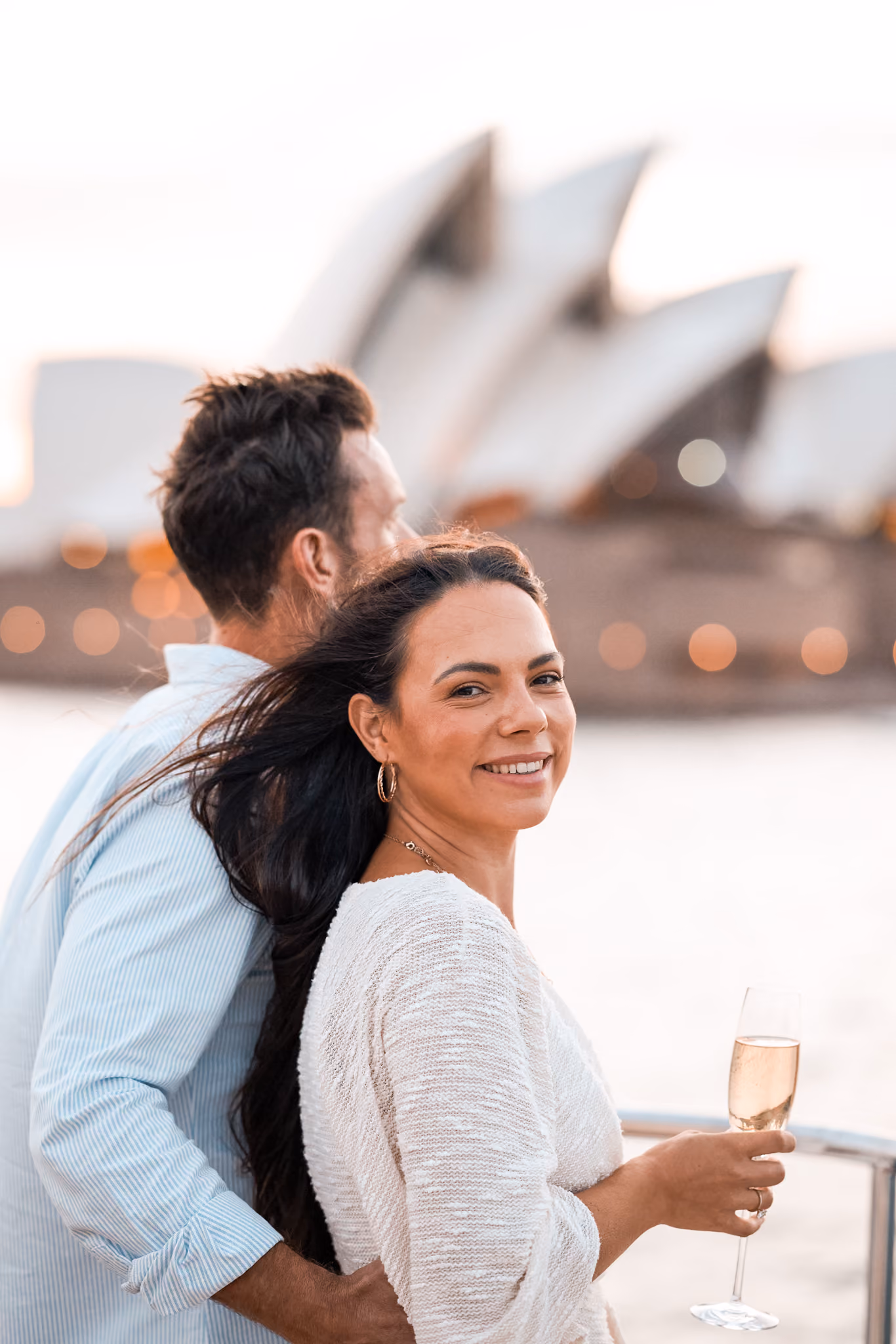 Smiling woman holding a glass of champagne with a man standing behind her near a railing, with the Sydney Opera House blurred in the background.