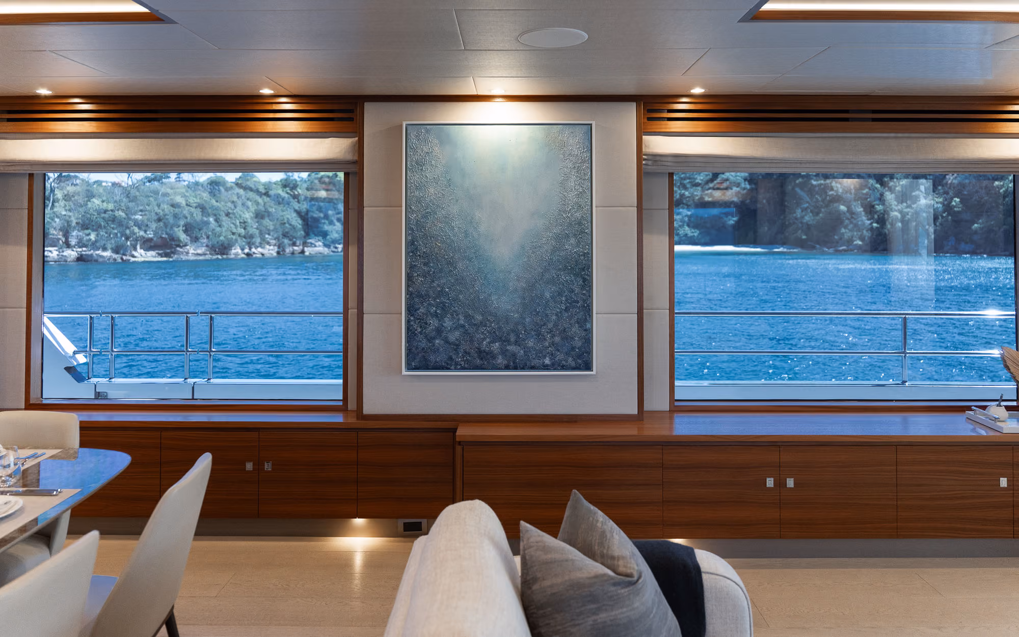Interior of a luxurious yacht dining and seating area with large windows revealing a calm blue sea and tree-covered coastline, featuring wooden accents and a modern abstract painting.