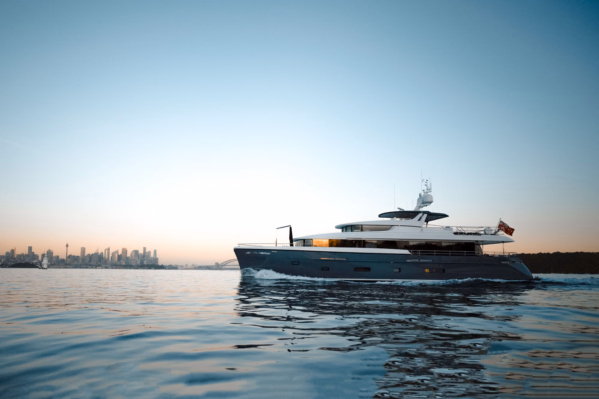 Belle, a luxury yacht cruising on calm water at sunset with Sydney skyline and Harbour Bridge in the distance.