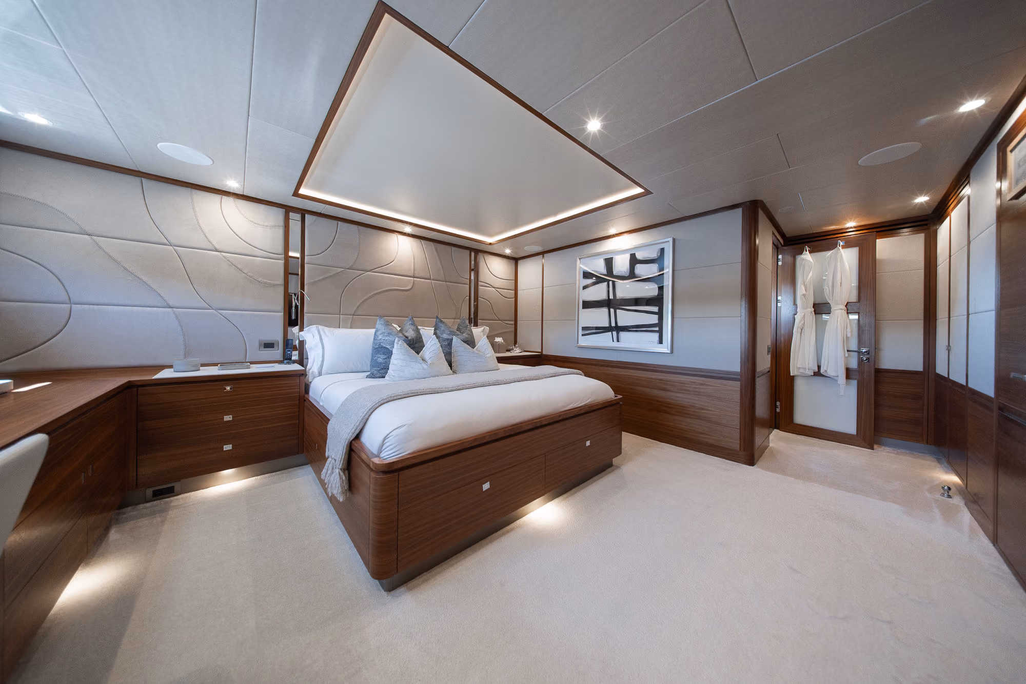 Modern, spacious bedroom with a large wooden-framed bed, white bedding and gray pillows, built-in wooden furniture, soft carpet, recessed lighting, an abstract black and white painting on the wall, and two white robes hanging on a door.