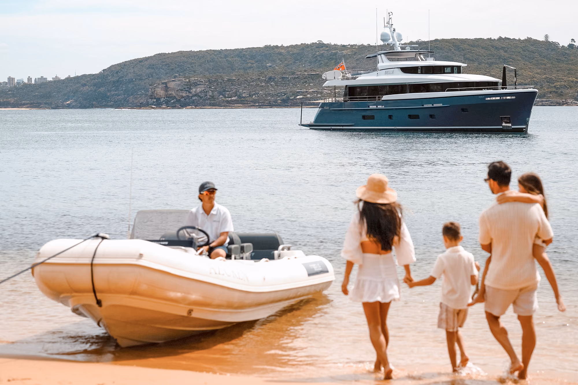 Family of four walking into shallow water toward a docked white inflatable boat with a man sitting at the helm, with a large yacht anchored further out on the water near a tree-covered hill.