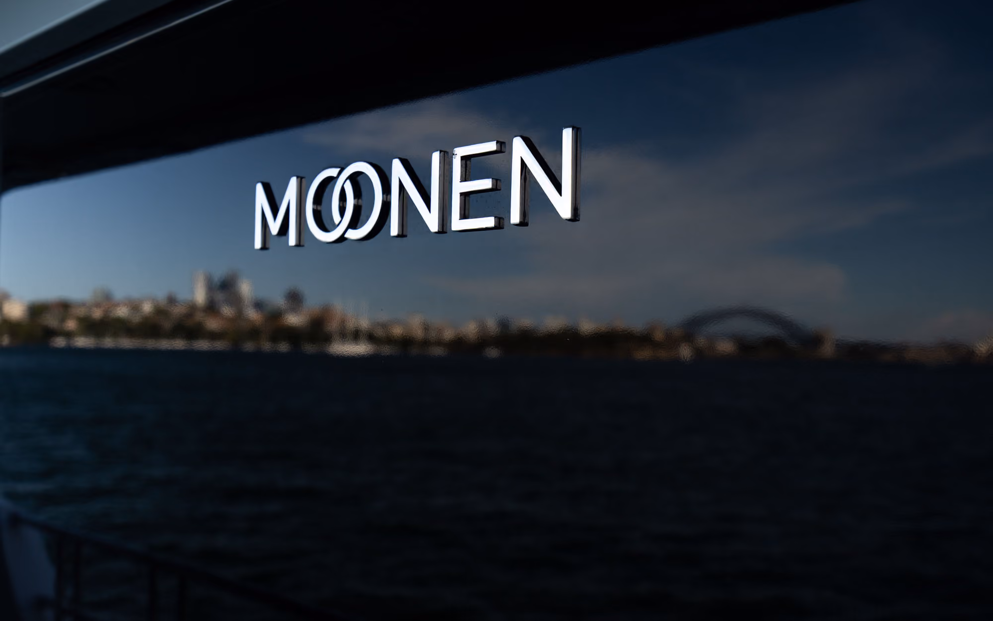 Reflective chrome text reading 'MOONEN' on a dark surface with an intertwined double 'O', with a blurred waterfront cityscape and a large bridge in the background under a blue sky.
