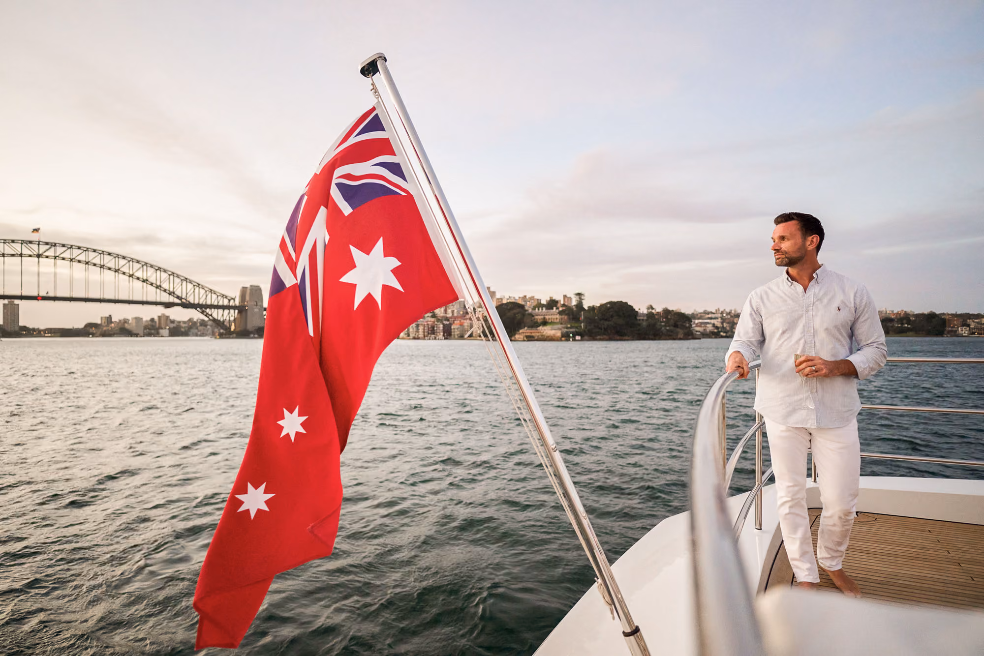 Man dressed in white standing barefoot on a boat deck holding the railing, with a red Australian flag flying on a pole and a city skyline including a bridge in the background over water.