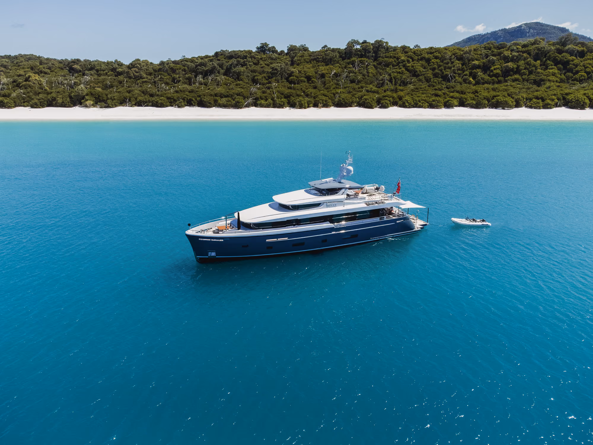 Luxury motor yacht anchored in clear turquoise water near a white sandy beach with dense green forest and hills in the background, accompanied by a small tender boat.