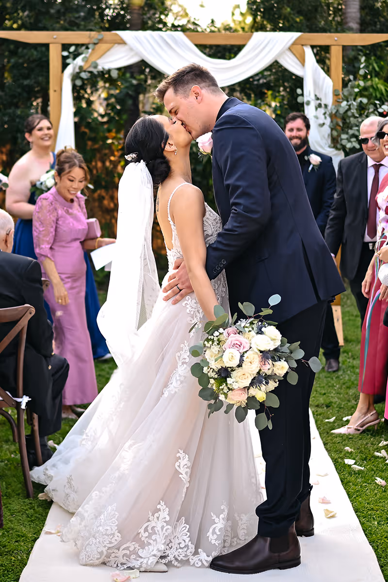 A couple kiss at their wedding ceremony as guests look on.