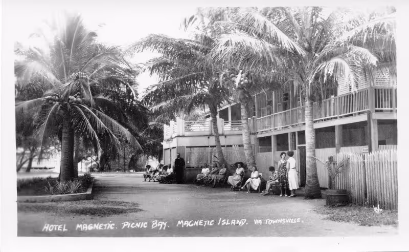A historic photograph of the original Picnic Bay Hotel, guests gathered under the palms out front.