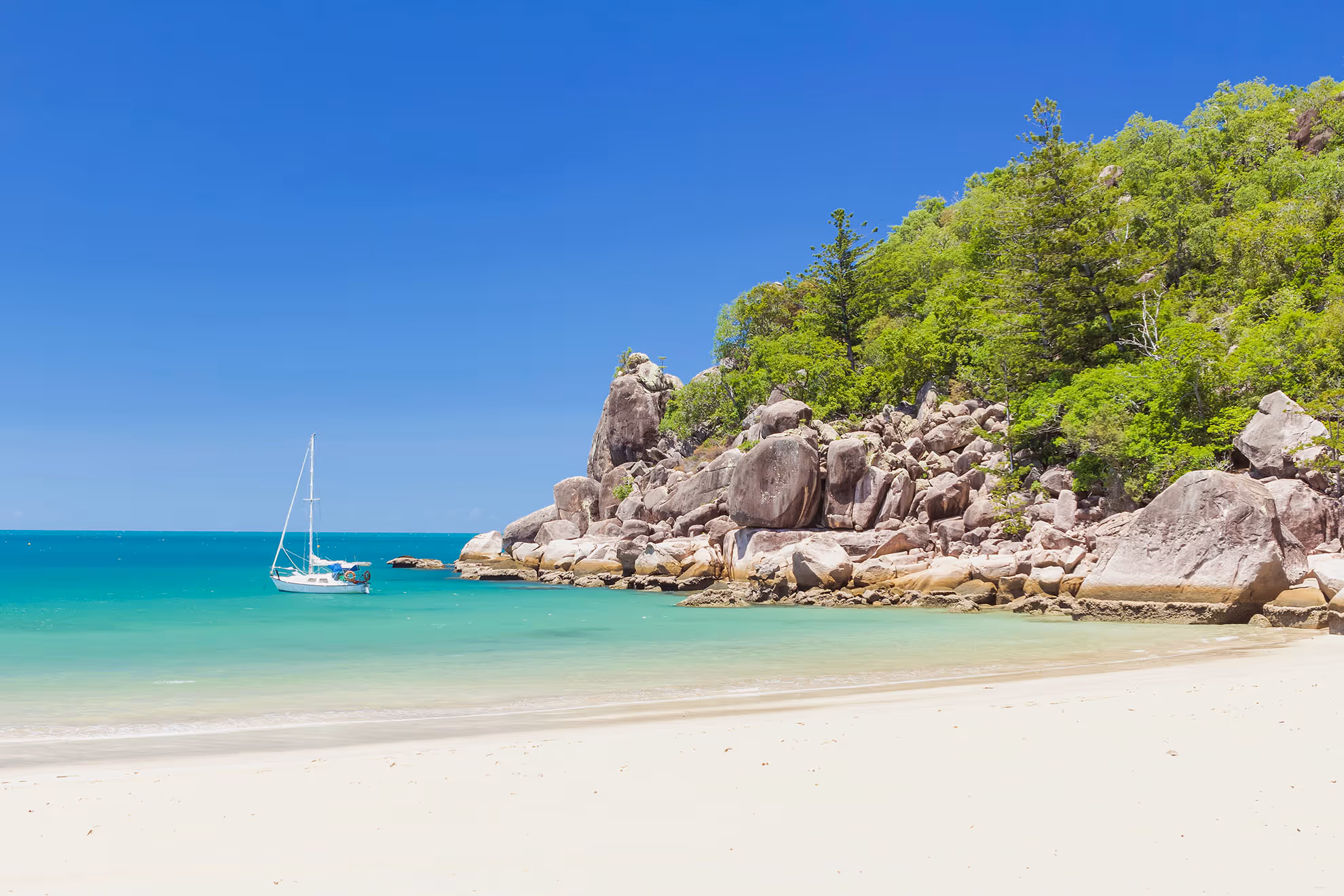A yacht at anchor off a quiet Magnetic Island beach scattered with granite boulders.