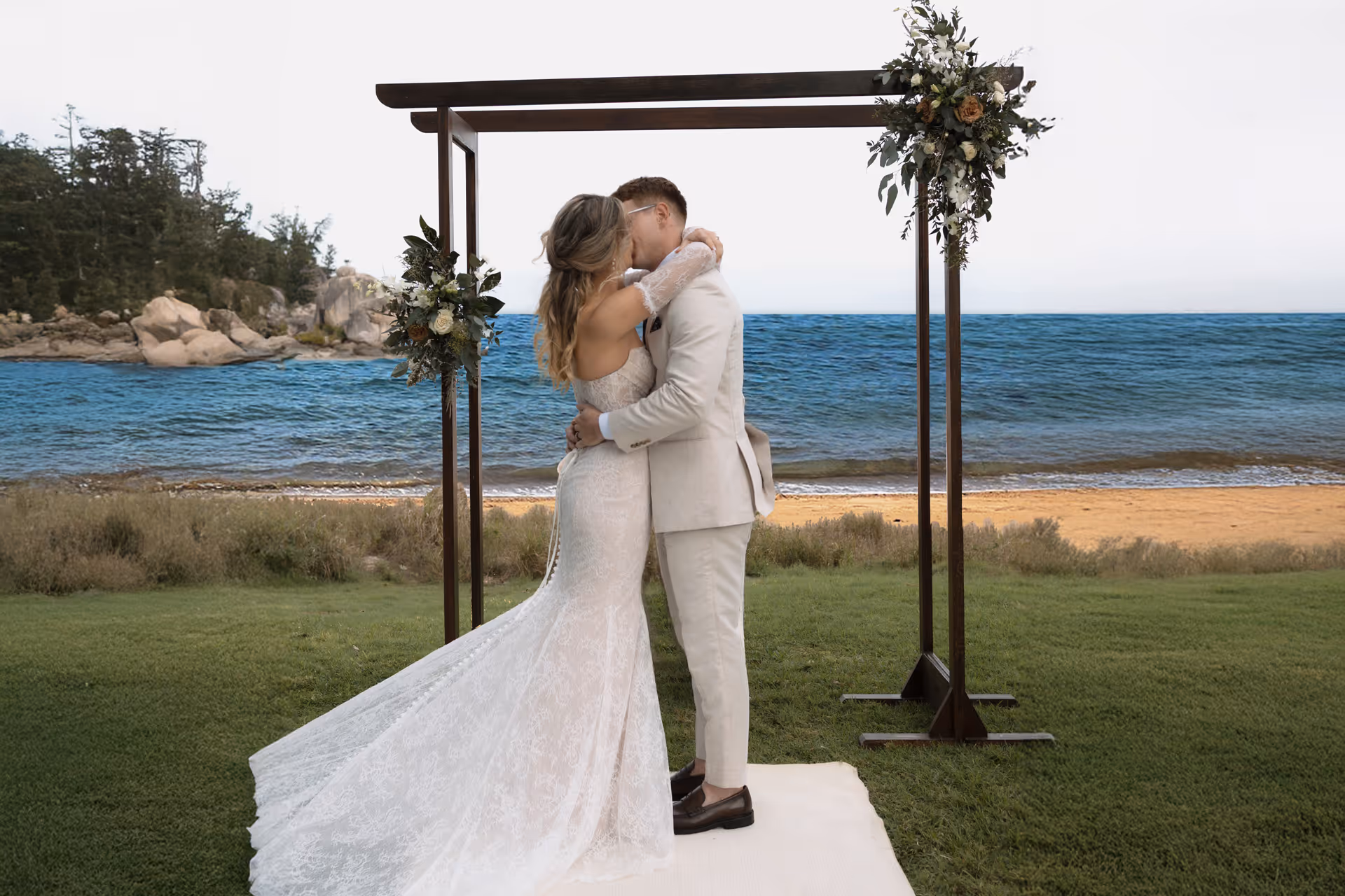 A couple embrace beneath a floral arbour on the lawn, the beach behind them.