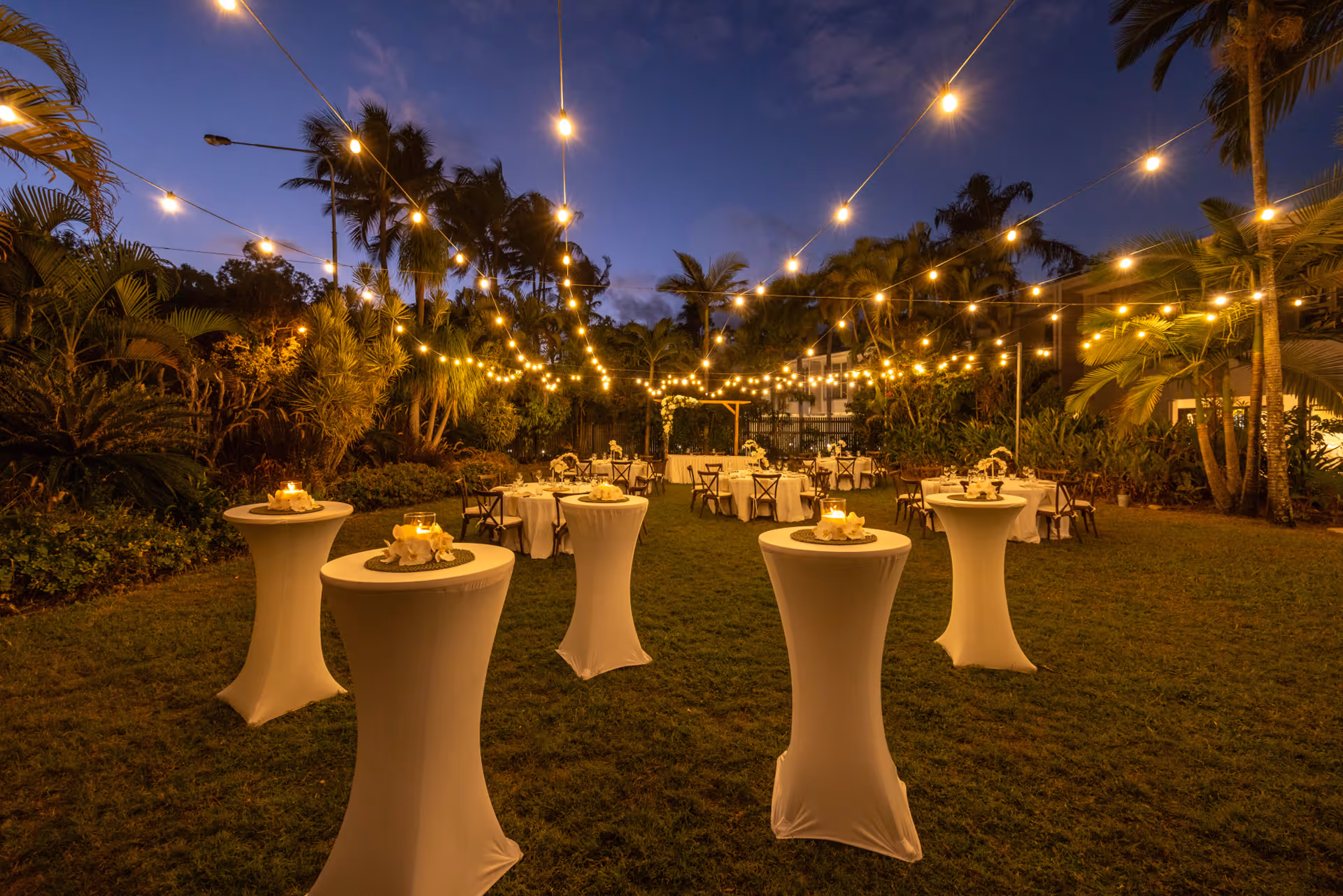 Cocktail tables set out on the lawn at dusk beneath strings of festoon lights.