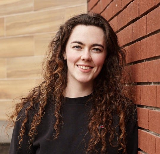 Smiling young woman with long curly dark brown hair wearing a black sweatshirt, standing against a brick wall and light wood paneling.