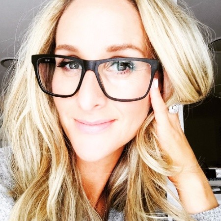 Close-up portrait of a smiling woman with long blonde hair wearing large black-framed glasses, resting her face on her hand showing a large ring.