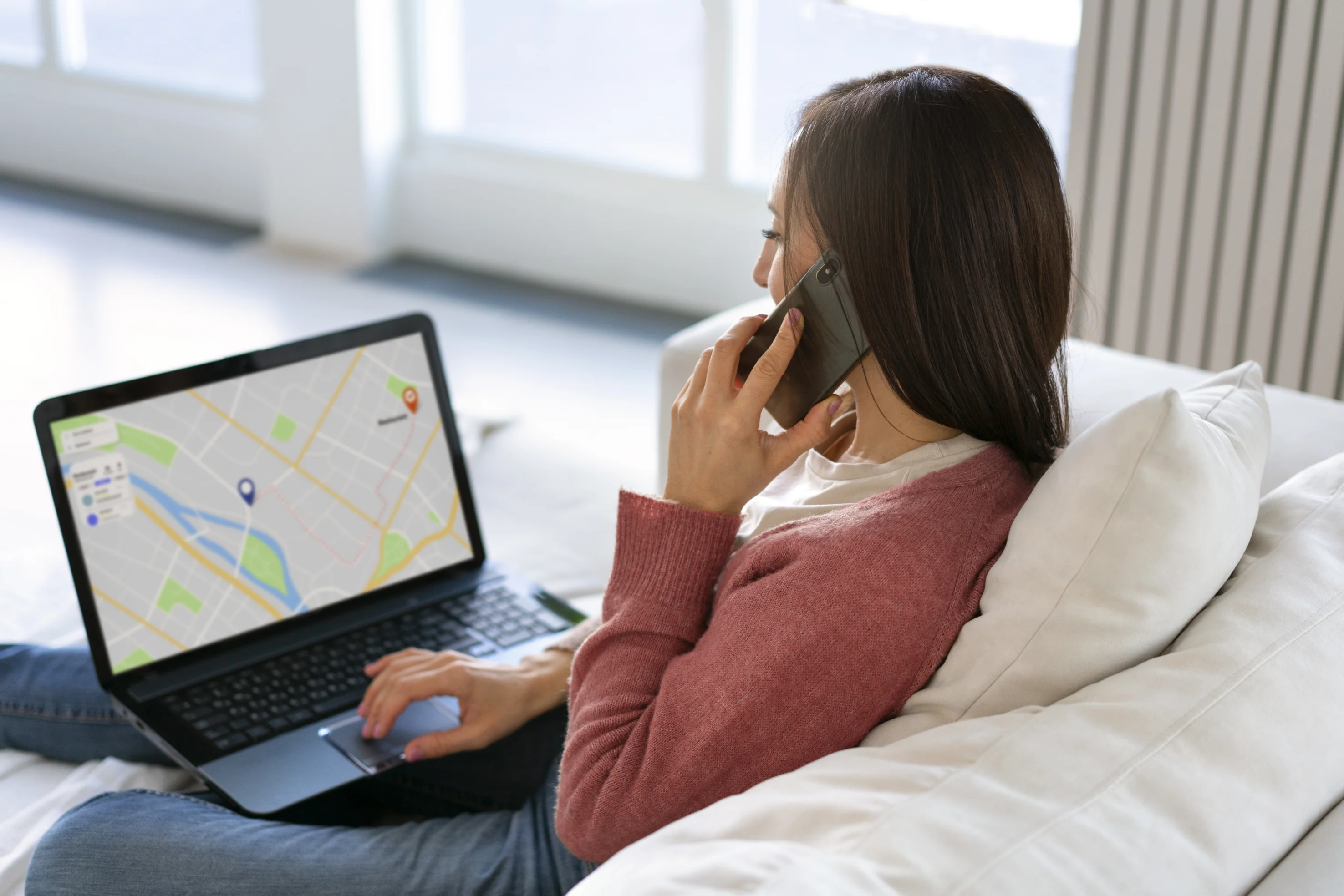 Woman sitting on a couch using a laptop displaying a map with two location markers while talking on a smartphone.
