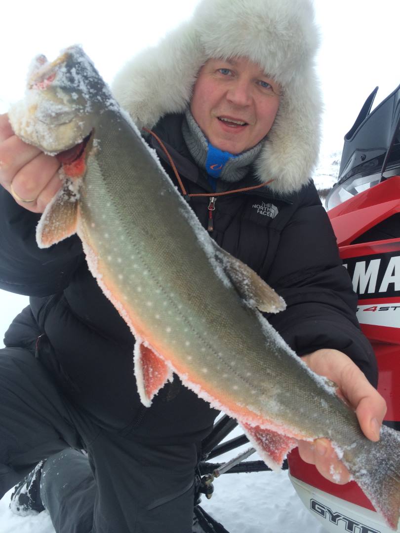 Mann iført tykk vinterjakke og pelslue holder en stor isbelagt fisk med oransje finner under isfiske i snødekt landskap.