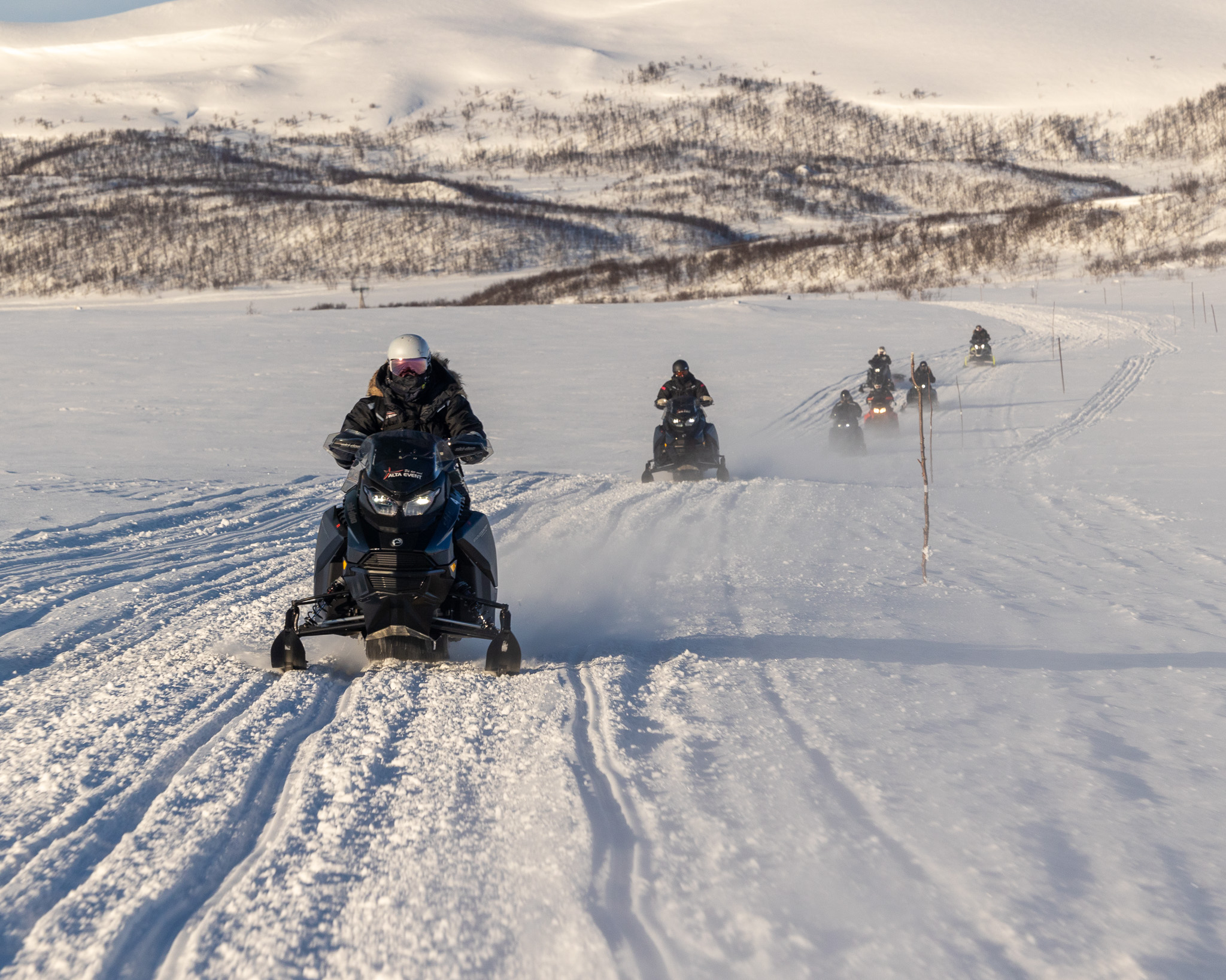 A group of snowmobiles driving through a snowy landscape with snow-covered mountains in the background under a clear sky.