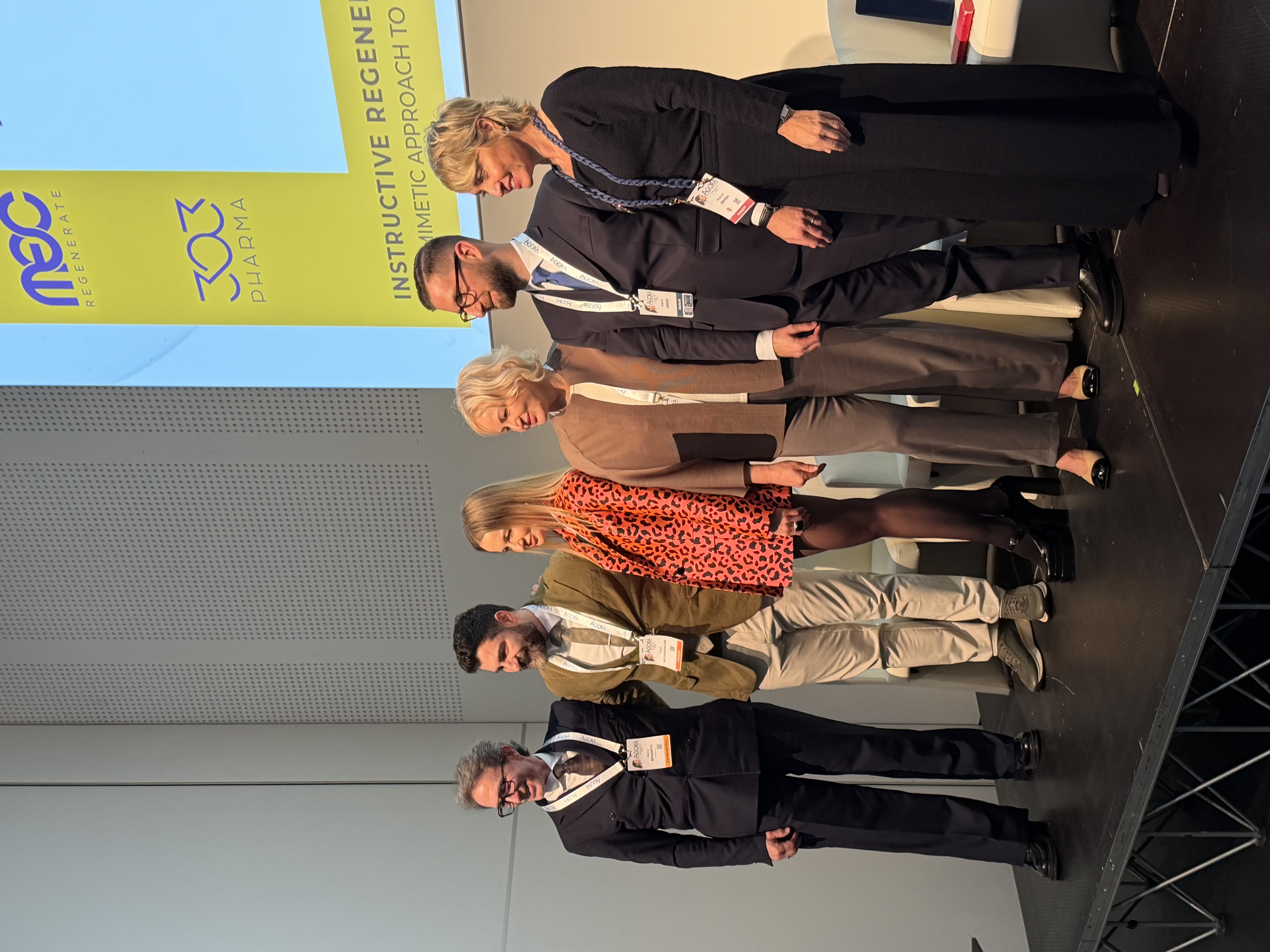 Dr Cemal with five other workshop participants on stage at Agorà 2026, with MEC Regenerate and 303 Pharma signage behind them.