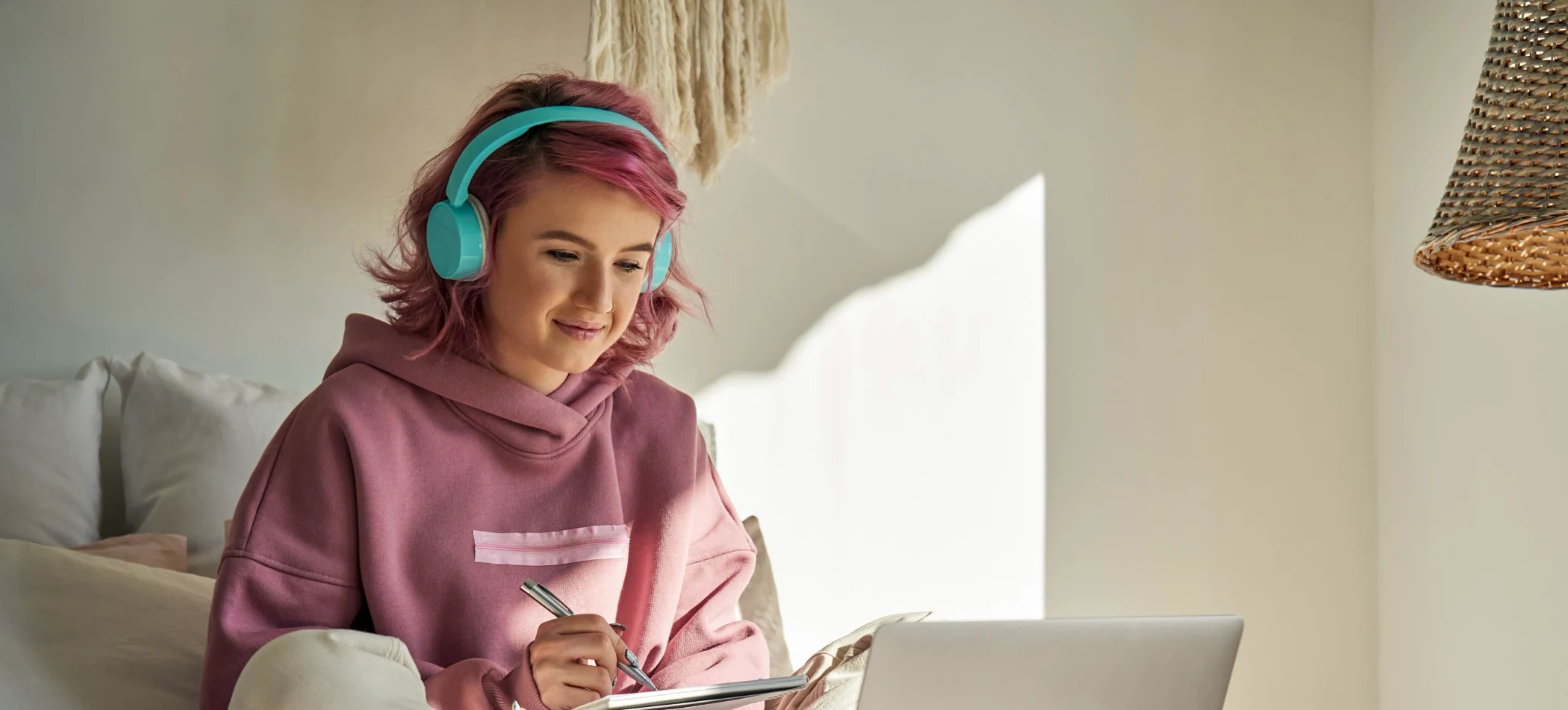 Young woman with pink hair wearing teal headphones and a pink hoodie sitting on a couch and writing on a notepad while looking at an open laptop.