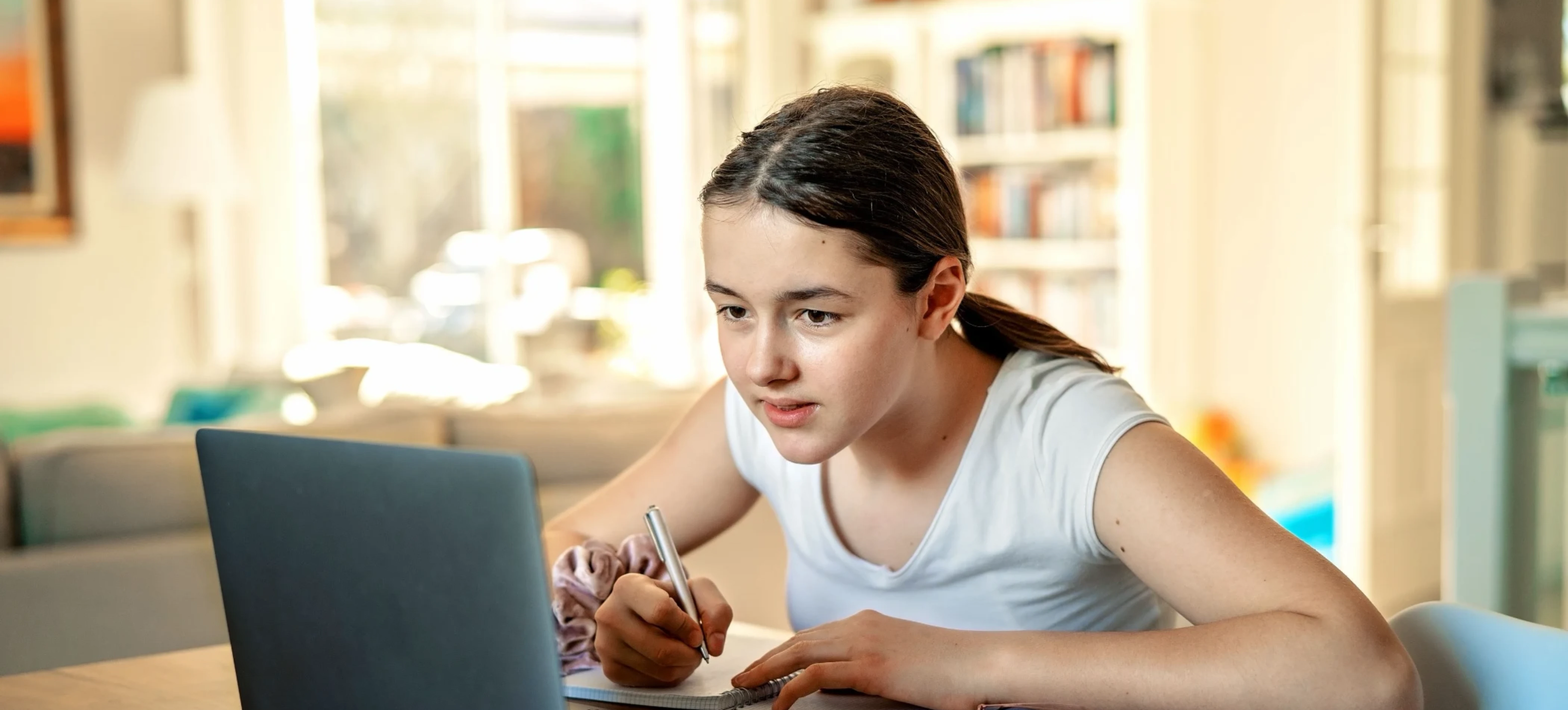 A young woman in a white shirt focused on her laptop while writing notes with a pen in a notebook at a table in a bright room.