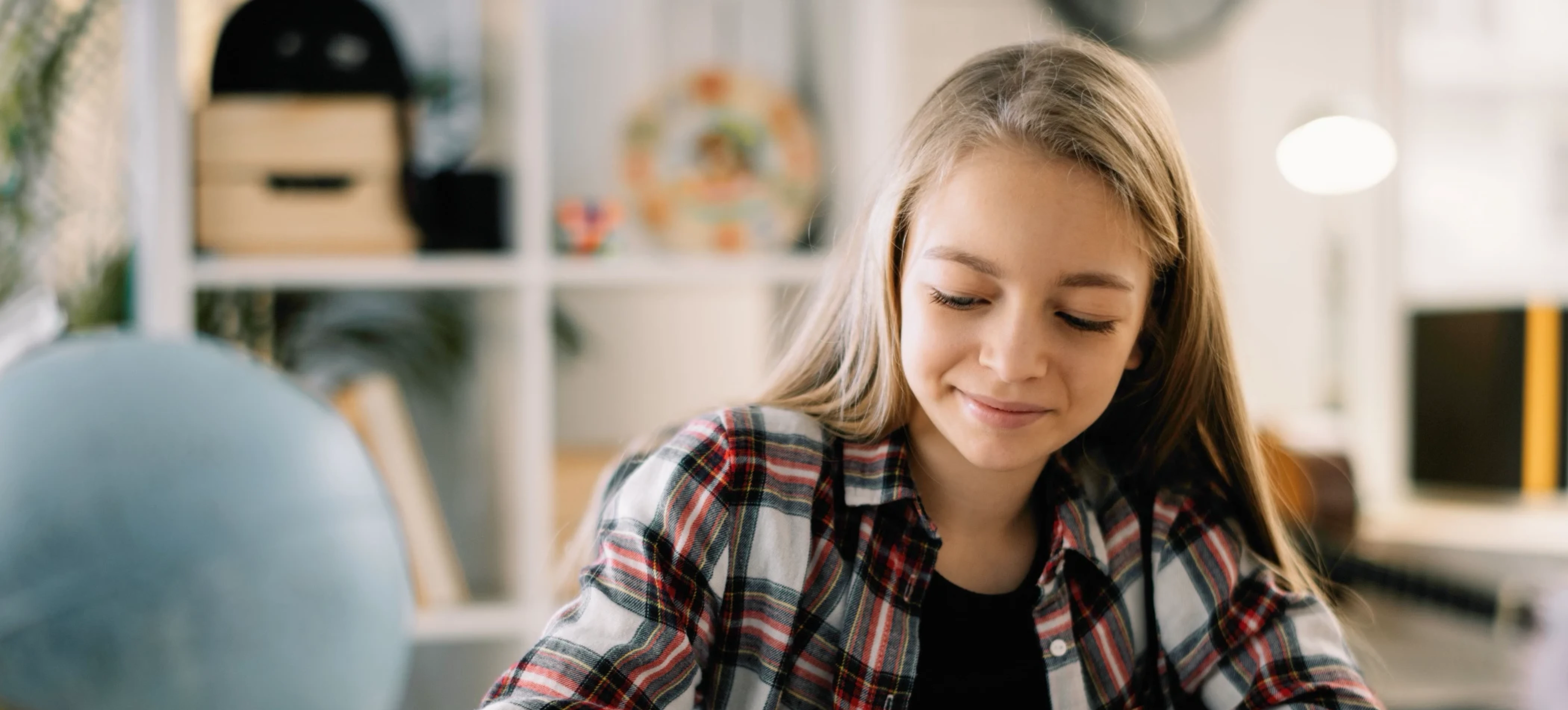Young girl with long blonde hair wearing a red and black plaid shirt, smiling slightly and looking down while seated indoors with a globe and bookshelf in the background.