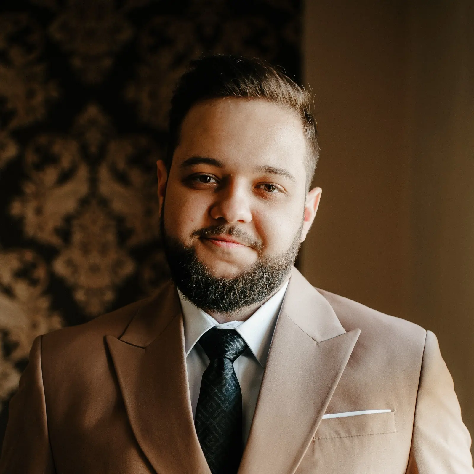 Portrait of a man with short hair and a beard, wearing a beige suit jacket, white shirt, and black patterned tie, smiling slightly in front of a dark ornate background.