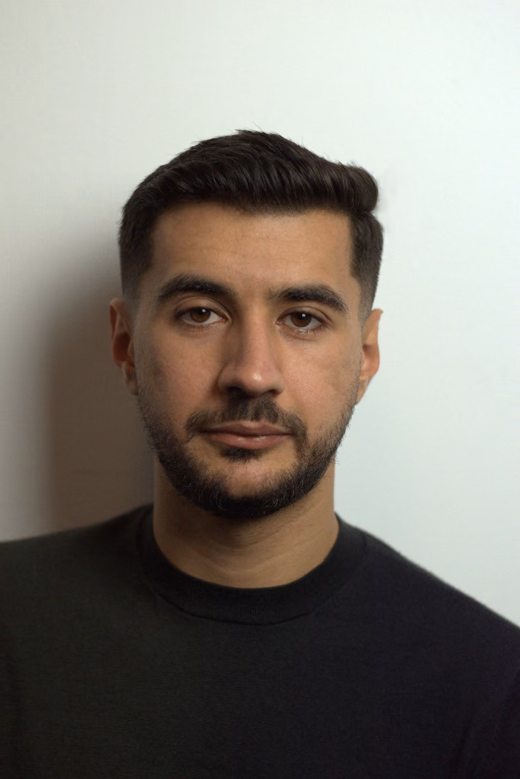 Portrait of a young man with short dark hair and a trimmed beard, wearing a black shirt, against a plain light background.