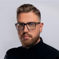 Portrait of a man with a beard and mustache wearing black-framed glasses and a black shirt against a plain light gray background.