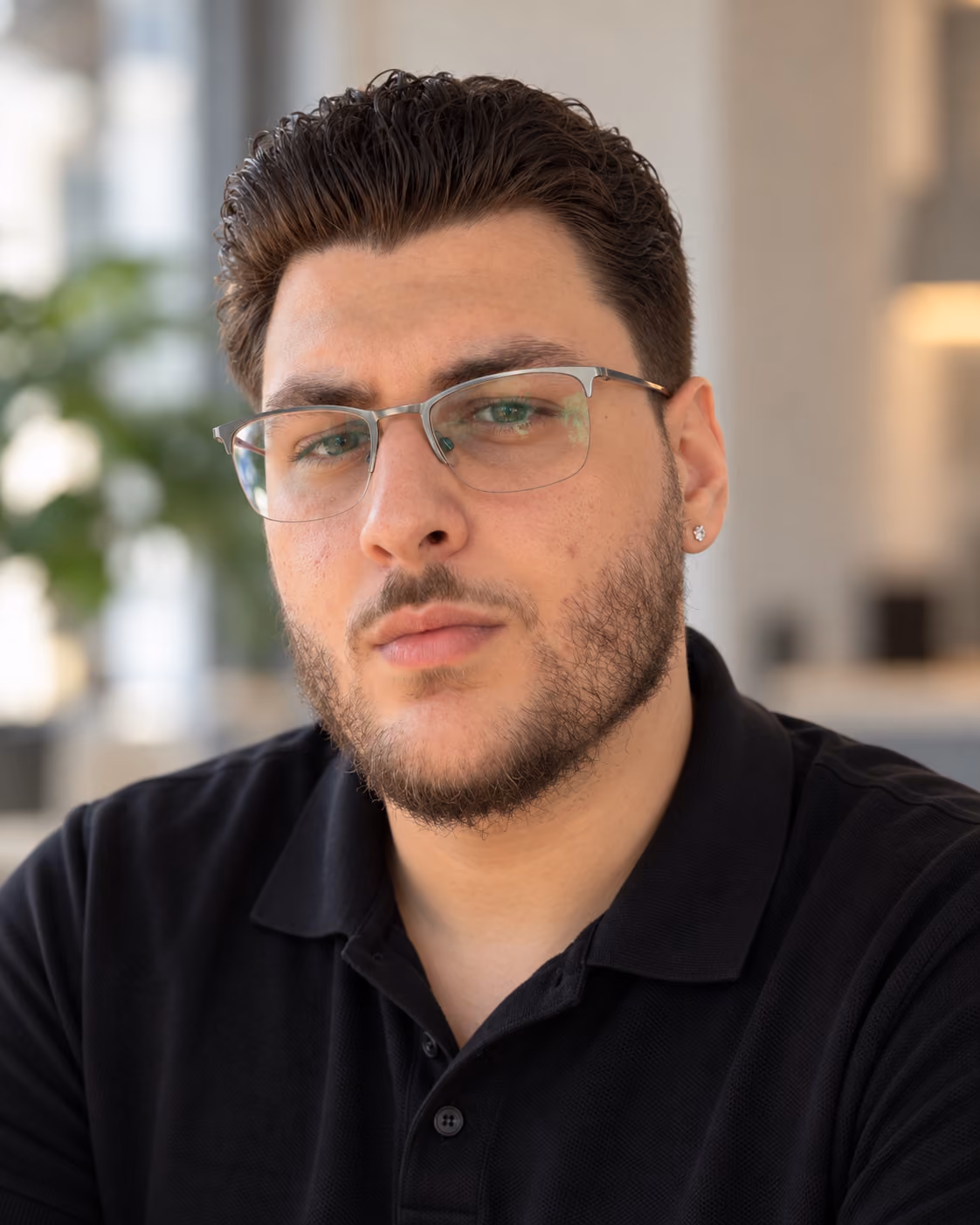 Portrait of a man with short dark hair, trimmed beard, glasses, and a black polo shirt in a softly lit indoor setting.