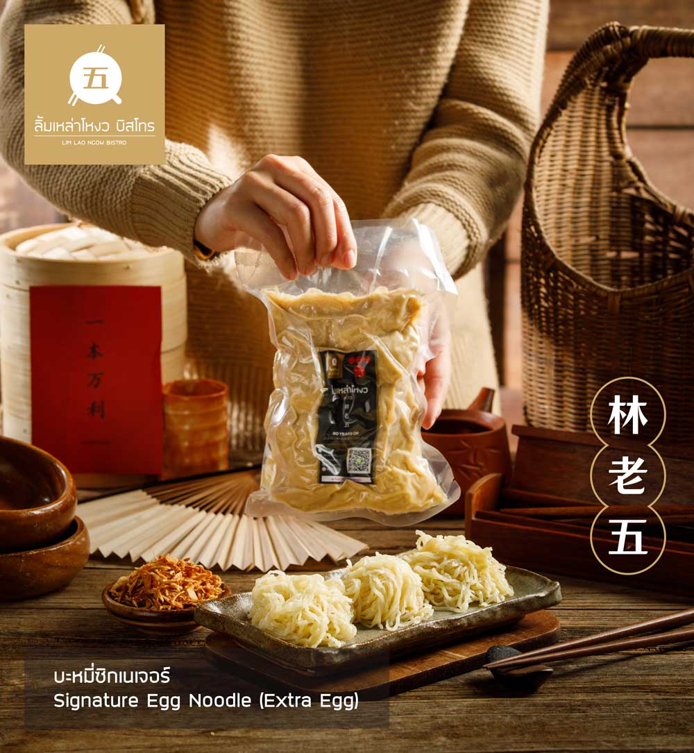 มือถือถุงบะหมี่ซิกเนเจอร์บะหมี่ไข่พิเศษ วางอยู่บนโต๊ะไม้ พร้อมจานบรรจุบะหมี่ไข่สามก้อน ข้างๆ มีชามไม้ ถ้วยเครื่องเคียง และตะเกียบ ด้านหลังมีของตกแต่งสไตล์เอเชีย พร้อมโลโก้ร้านลิ้มเหล่าโหงว บิสโทร