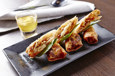 Spring rolls sliced in half showing crispy fried exterior and filling, served on a black rectangular plate with green leaves inside and a small glass of yellow dipping sauce on the side.