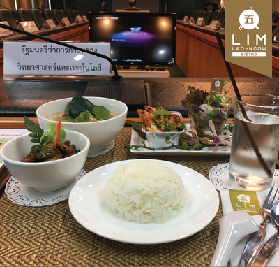 ชุดอาหารไทยในร้านอาหาร LIM Lao-Ngow Bistro ประกอบด้วยข้าวสวย ต้มยำ ตำรับแกงถ้วย และน้ำดื่มวางบนโต๊ะที่มีผ้าปูโต๊ะลายสานพร้อมถ้วยชามช้อนส้อมและแก้วน้ำใส ใต้โต๊ะมีจานรองและในแบ็คกราวด์มีหน้าจอทีวีและป้ายที่เขียนว่า รัฐมนตรีว่าการกระทรวงวิทยาศาสตร์และเทคโนโลยี