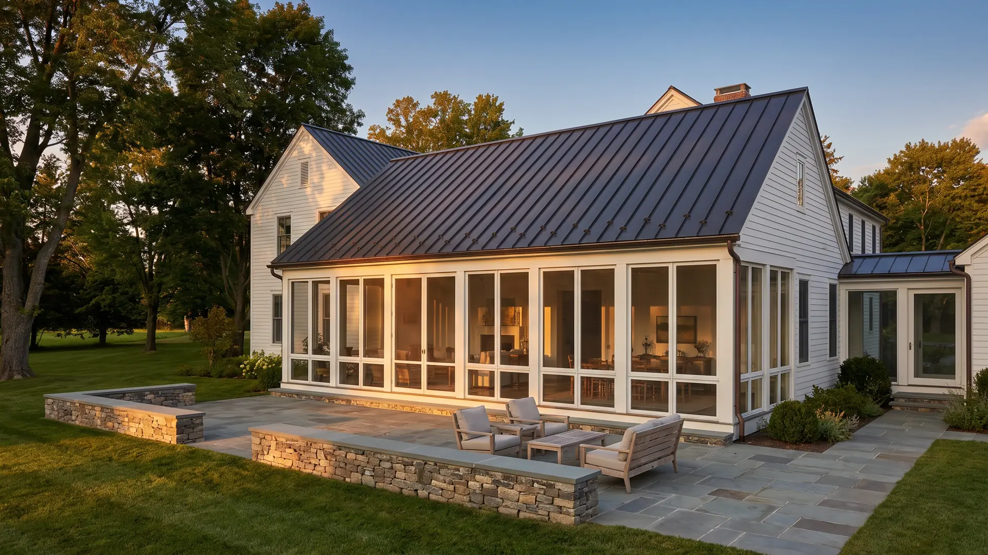 A white farmhouse with a glass garden-room wing and a bluestone terrace at golden hour