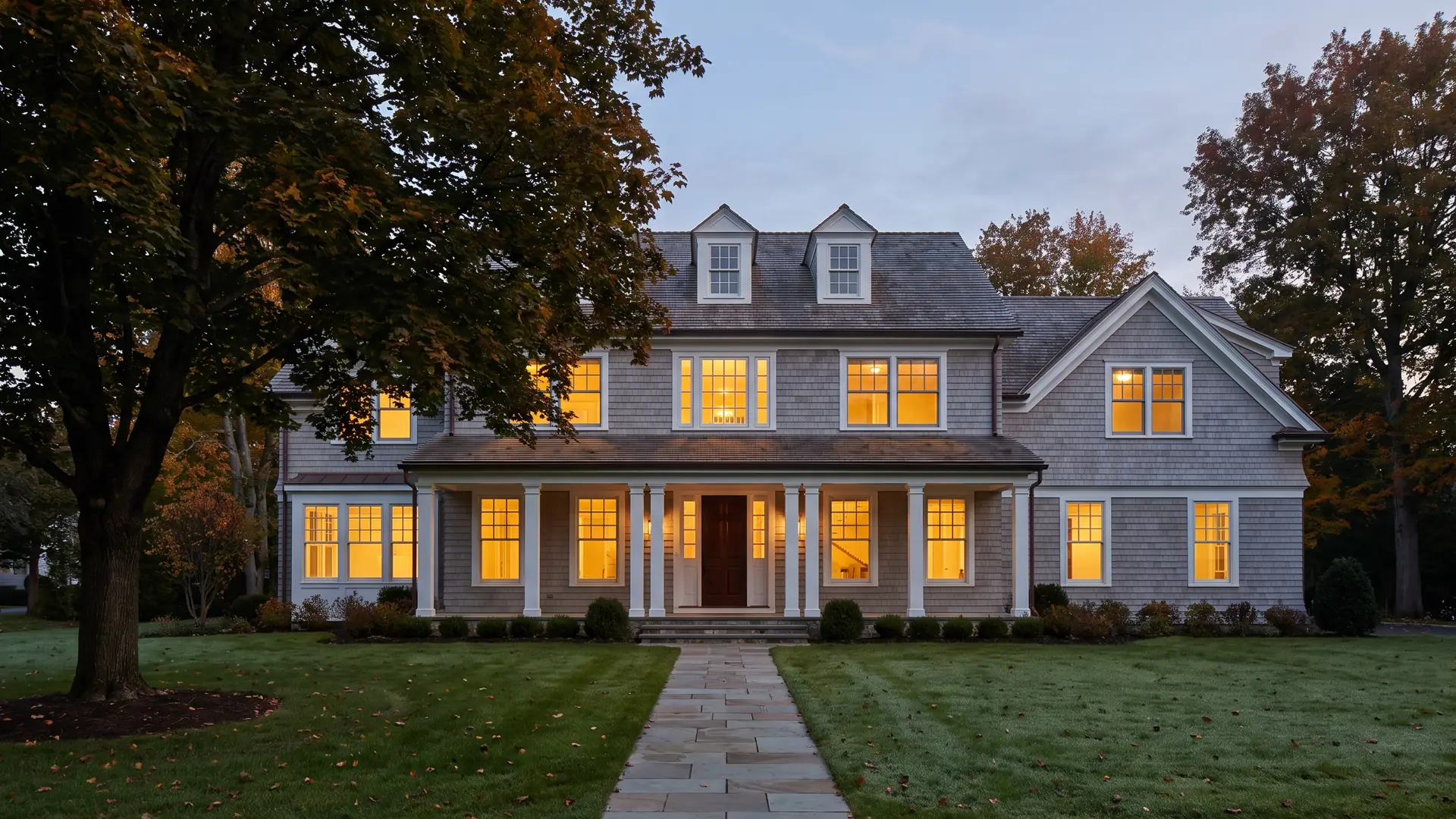 A gray shingle-style colonial at dusk with the windows lit and a bluestone walk