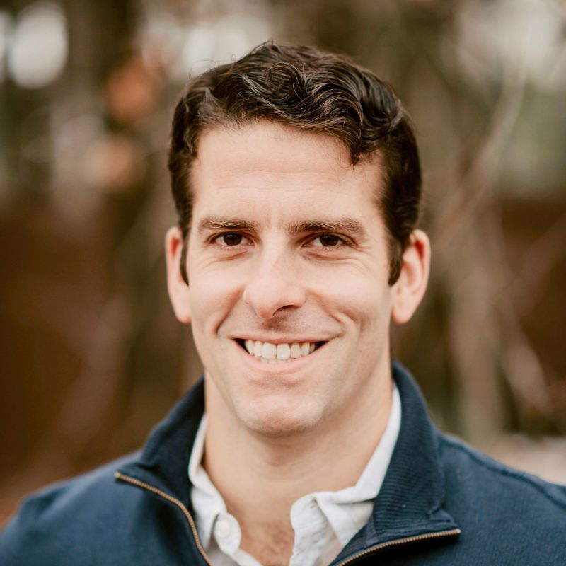Smiling man with short, dark curly hair wearing a navy blue jacket over a white collared shirt against a blurred natural background.