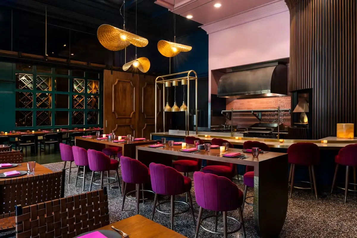 Modern restaurant interior with dark wood tables, magenta upholstered chairs, warm hanging lights, and a bar with candles.